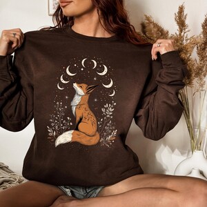 K&ouml;nnte beinhalten: Ein braunes Sweatshirt mit einer Fuchsgrafik und einem Mond- und Stern-Design. Der Fuchs ist in Orange und Wei&szlig; gehalten und von floralen Elementen umgeben. Das Sweatshirt hat lange &Auml;rmel und einen Rundhalsausschnitt.