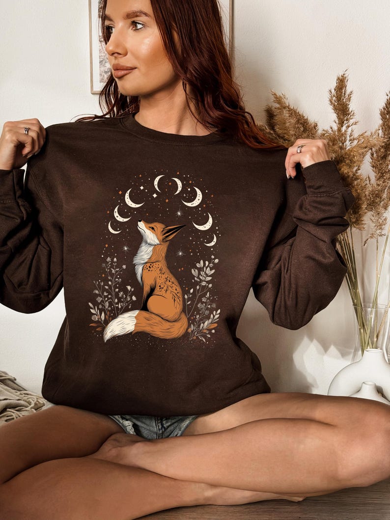 K&ouml;nnte beinhalten: Ein braunes Sweatshirt mit einer Fuchsgrafik und einem Mond- und Stern-Design. Der Fuchs ist in Orange und Wei&szlig; gehalten und von floralen Elementen umgeben. Das Sweatshirt hat lange &Auml;rmel und einen Rundhalsausschnitt.