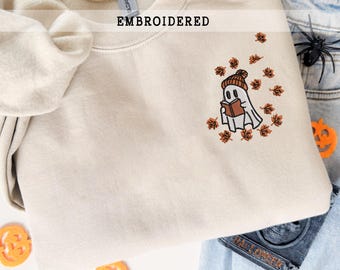 May include: A beige crewneck sweatshirt featuring an embroidered design of a white ghost wearing an orange beanie and reading a brown book, surrounded by small orange and brown autumn leaves. The word EMBROIDERED is printed above the design.