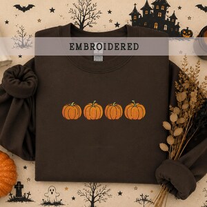 May include: A dark brown crewneck sweatshirt featuring an embroidered design of four orange pumpkins arranged in a horizontal row across the center of the chest. The garment has long sleeves with ribbed cuffs and a ribbed neckline.