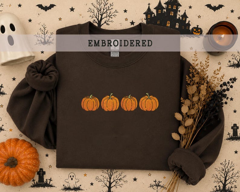 May include: A dark brown crewneck sweatshirt featuring an embroidered design of four orange pumpkins arranged in a horizontal row across the center of the chest. The garment has long sleeves with ribbed cuffs and a ribbed neckline.