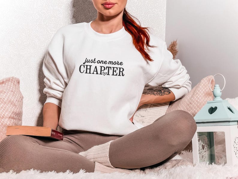 Pode incluir: Camisola branca com o texto preto "just one more CHAPTER" e um s&iacute;mbolo de cora&ccedil;&atilde;o. A pessoa est&aacute; sentada com um livro no colo. A pessoa est&aacute; usando leggings marrons e meias brancas.