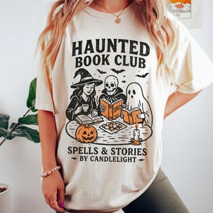May include: Cream-colored t-shirt with a Halloween-themed graphic. The design features the text "HAUNTED BOOK CLUB" above illustrations of a witch, a skeleton, and a ghost reading books. Below, it reads "SPELLS & STORIES BY CANDLELIGHT".