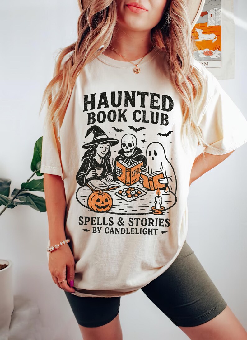 May include: Cream-colored t-shirt with a Halloween-themed graphic. The design features the text "HAUNTED BOOK CLUB" above illustrations of a witch, a skeleton, and a ghost reading books. Below, it reads "SPELLS & STORIES BY CANDLELIGHT".