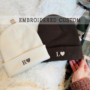Peut inclure: Deux bonnets personnalisés brodés. L'un est de couleur crème avec "R" et un cœur, l'autre est marron foncé avec "L" et un cœur. Le texte "EMBROIDERED CUSTOM" est au-dessus des bonnets. Les bonnets sont sur une surface blanche duveteuse.