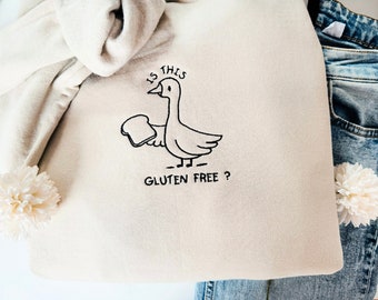 May include: A beige tote bag with a black embroidered design of a goose holding a slice of bread. The text "Is this gluten free?" is embroidered below the goose.