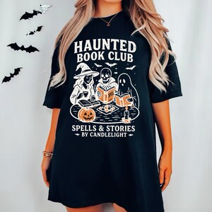 May include: Black t-shirt featuring a Halloween-themed graphic. The design includes the text "HAUNTED BOOK CLUB" above an illustration of a witch, a skeleton, and a ghost reading books. The phrase "SPELLS & STORIES BY CANDLELIGHT" is below the illustration.