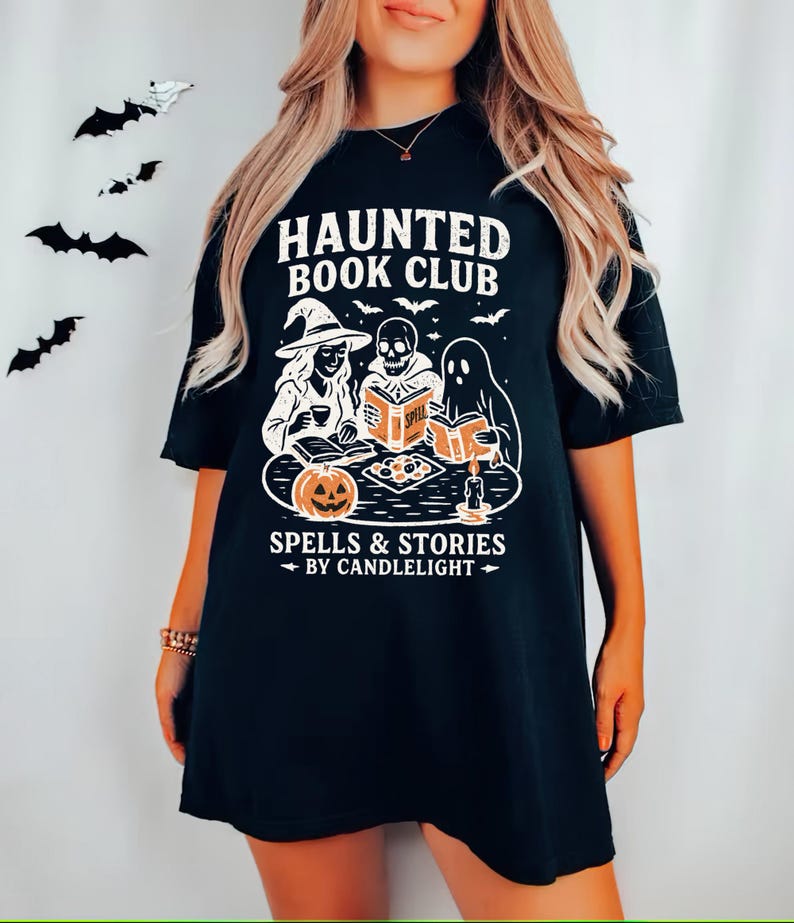 May include: Black t-shirt featuring a Halloween-themed graphic. The design includes the text "HAUNTED BOOK CLUB" above an illustration of a witch, a skeleton, and a ghost reading books. The phrase "SPELLS & STORIES BY CANDLELIGHT" is below the illustration.
