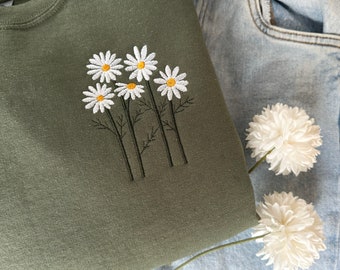 Könnte beinhalten: Ein grüner Pullover mit gestickten weißen Gänseblümchen auf der Vorderseite. Die Gänseblümchen haben gelbe Zentren und sind in einer Gruppe angeordnet.