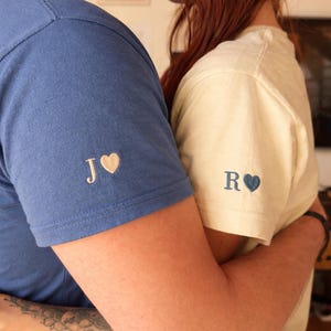 Puede incluir: Dos personas abrazándose, vistiendo camisetas a juego. Una es azul con "J" y un corazón, la otra crema con "R" y un corazón. Las camisetas están personalizadas con iniciales bordadas y símbolos de corazón.