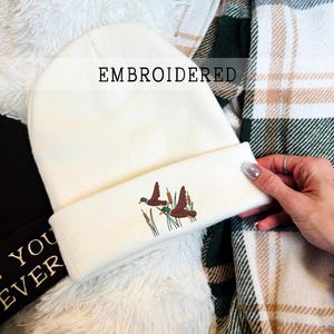 Op de afbeelding: Crèmekleurige muts met een geborduurd ontwerp van twee eenden in vlucht boven riet. Het woord "EMBROIDERED" staat op de hoed. De hoed is omgeslagen aan de rand en ligt op een witte, pluizige ondergrond.