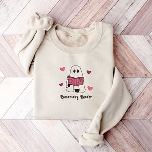 May include: A cream-colored sweatshirt with a graphic of a ghost reading a book with pink hearts around it. The text "Romanstasy Reader" is printed below the ghost.