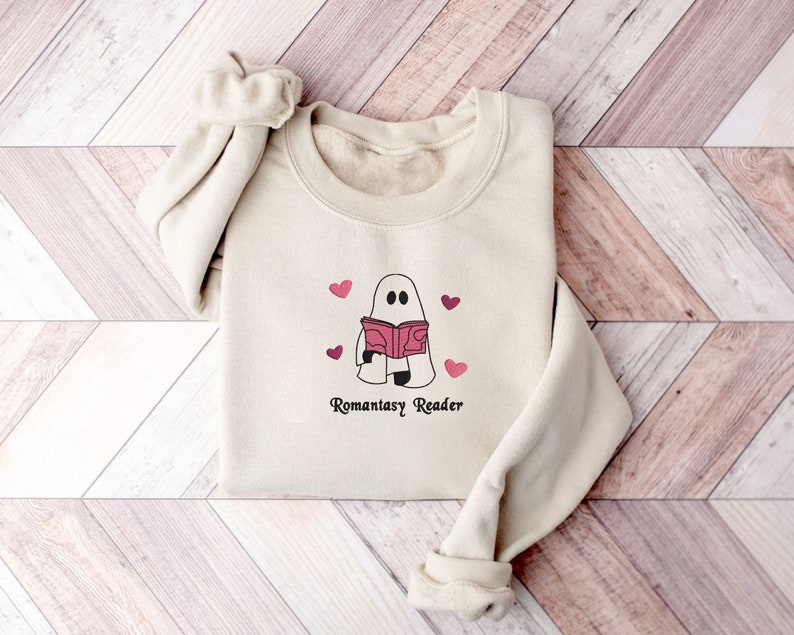 May include: A cream-colored sweatshirt with a graphic of a ghost reading a book with pink hearts around it. The text "Romanstasy Reader" is printed below the ghost.
