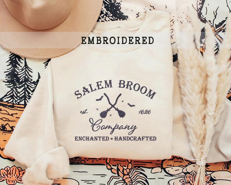 K&ouml;nnte beinhalten: Ein cremefarbener Pullover mit dem Text "Salem Broom Company", der in Schwarz gestickt ist. Der Text ist von einem Kreis mit den Worten "est. 1626" und zwei gekreuzten Besen umgeben.