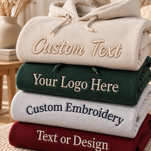 Könnte beinhalten: Ein Stapel aus vier gefalteten Kapuzenpullovern in Beige, Dunkelgrün, Hellgrau und Kastanienbraun. Jeder Pullover hat eine mittige Stickerei. Der beige Pullover zeigt Custom Text, der grüne Your Logo Here, der graue Custom Embroidery und der kastanienbraune Text or Design.