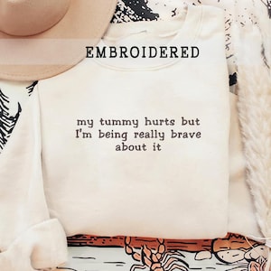 Puede incluir: Una sudadera blanca con el texto "my tummy hurts but I'm being really brave about it" bordado en la parte delantera con hilo marrón.