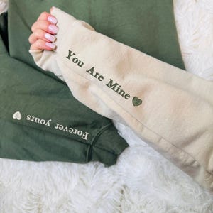 Op de afbeelding: Twee sweatshirts, één olijfgroen en één beige, met de woorden "Forever Yours" en "You Are Mine" op de mouwen. Elke mouw heeft een kleine hartafbeelding. De sweatshirts worden gepresenteerd op een zacht wit oppervlak.