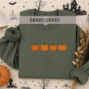 May include: An olive green crewneck sweatshirt featuring an embroidered design of four orange pumpkins in a horizontal row across the center. The garment has long sleeves with ribbed cuffs and a ribbed neckline, showcasing a clean, simple aesthetic.