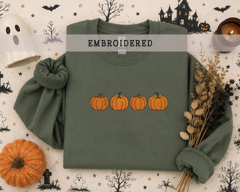 May include: An olive green crewneck sweatshirt featuring an embroidered design of four orange pumpkins in a horizontal row across the center. The garment has long sleeves with ribbed cuffs and a ribbed neckline, showcasing a clean, simple aesthetic.