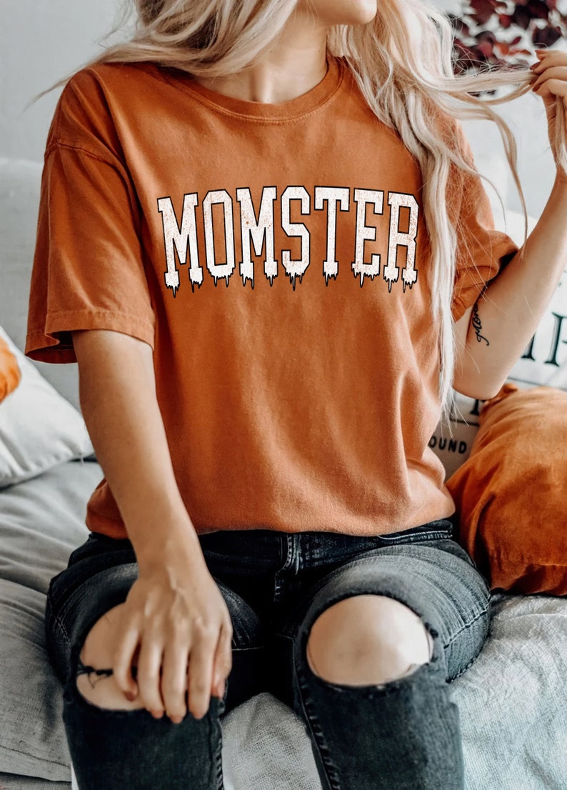 May include: A burnt orange t-shirt with the text "MOMSTER" in white dripping letters. The shirt is worn by a person sitting on a bed with a white blanket.