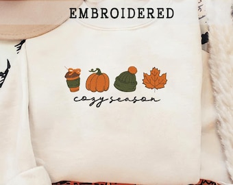 Könnte beinhalten: Cremefarbenes Sweatshirt mit dem Wort "EMBROIDERED" über gestickten Herbstmotiven: einer Kaffeetasse, einem Kürbis, einer Mütze und einem Ahornblatt. Darunter steht "cozy season".