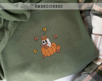 Puede incluir: Sudadera verde oliva con un diseño bordado. El diseño presenta un fantasma leyendo un libro sentado en una calabaza, rodeado de hojas de otoño. La palabra "EMBROIDERED" está impresa en la parte superior.