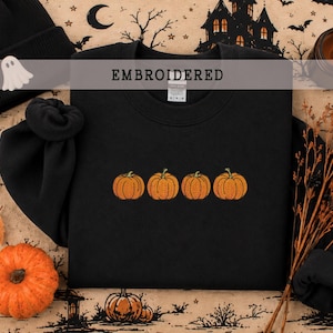 May include: A black crewneck sweatshirt featuring an embroidered design of four orange pumpkins in a horizontal row across the center. The garment has a ribbed collar, cuffs, and hem, with the word EMBROIDERED printed in white text above.