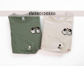 Könnte beinhalten: Zwei langärmlige Sweatshirts, eines olivgrün und eines cremefarben, werden gezeigt. Jedes hat ein gesticktes Design von zwei Pinguinen mit einem roten Herzen. Das Wort "EMBROIDERED" ist oben aufgedruckt. Der linke Ärmel jedes Shirts hat einen Buchstaben.
