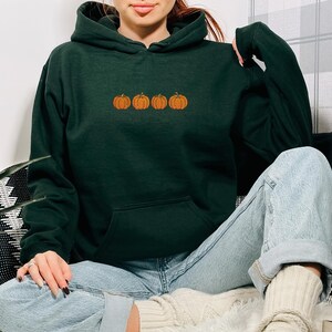 May include: A dark forest green hooded sweatshirt featuring a horizontal row of four small, embroidered orange pumpkins centered on the chest. The garment has long sleeves, a front kangaroo pocket, and a drawstring hood in the same dark green.