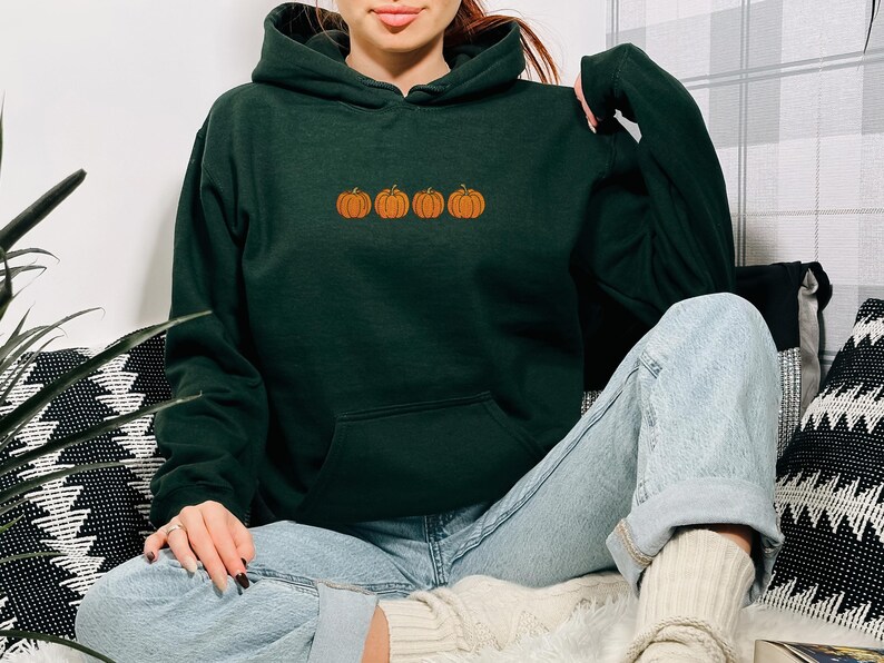 May include: A dark forest green hooded sweatshirt featuring a horizontal row of four small, embroidered orange pumpkins centered on the chest. The garment has long sleeves, a front kangaroo pocket, and a drawstring hood in the same dark green.