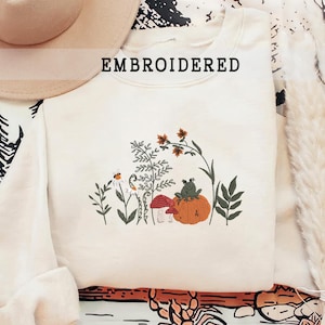 May include: Cream-colored embroidered sweatshirt with the word "EMBROIDERED" across the top. The design features a pumpkin, mushrooms, a frog, and various plants in shades of green, orange, and white.