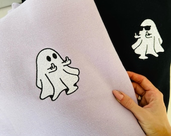 Puede incluir: Una sudadera de color morado claro con un gráfico bordado de un fantasma blanco con ojos negros y manos levantadas en un gesto obsceno, junto a una sudadera negra con un gráfico de un fantasma blanco con gafas de sol negras.