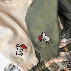 May include: Close-up of two sweatshirts, one cream and one olive green, with a duck graphic holding a red heart on the sleeves. A plaid flannel shirt is partially visible in the background.