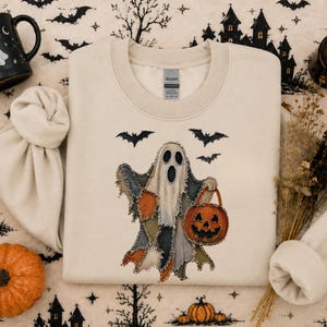 Peut inclure: Un sweat-shirt à col rond de couleur crème arborant un motif central représentant un fantôme composé de divers morceaux de tissu, tenant un seau citrouille orange. Trois chauves-souris noires sont placées au-dessus du fantôme, avec un aspect effiloché.