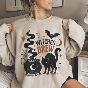 May include: A beige crewneck sweatshirt featuring a black and orange graphic print. The design includes a cauldron with rising smoke, a black cat, a bat, a spider web, stars, a crescent moon, and the text "WITCHES BREW" in a bold font.
