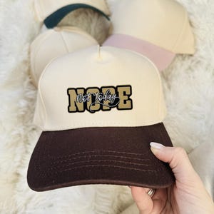 Op de afbeelding: Een verzameling baseballpetten in verschillende kleuren. De voorste pet is crèmekleurig met een bruin vizier en de tekst "Nope Not Today" in goud en zwart. Andere petten hebben teal en roze accenten.