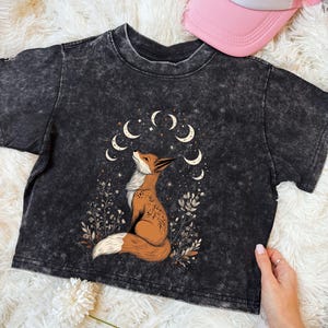 May include: A black acid wash crop top featuring a detailed illustration of a fox gazing upwards, surrounded by crescent moons and foliage. The fox is brown and white. A pink and white trucker hat is visible in the background.