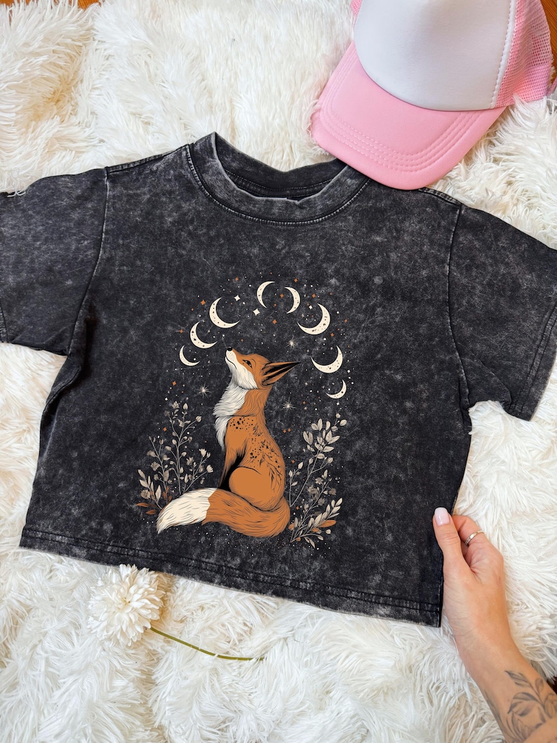 May include: A black acid wash crop top featuring a detailed illustration of a fox gazing upwards, surrounded by crescent moons and foliage. The fox is brown and white. A pink and white trucker hat is visible in the background.