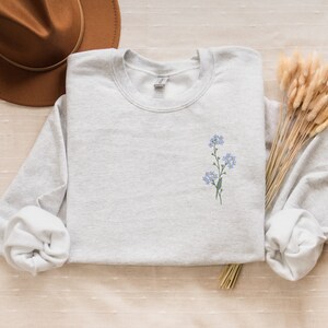 May include: A light gray sweatshirt with a small embroidered design of blue flowers on the left chest.