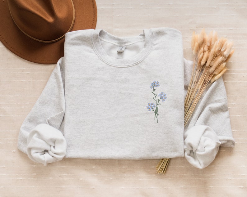 May include: A light gray sweatshirt with a small embroidered design of blue flowers on the left chest.