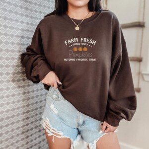 Op de afbeelding: Een donkerbruine sweatshirt met de witte tekst "Farm Fresh Picked Daily Pumpkins Autumns Favorite Treat". Het ontwerp bevat drie oranje pompoenen. De sweatshirt wordt gedragen met een spijkerbroekshort.