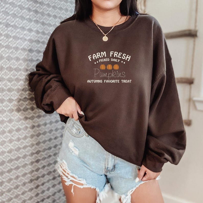 Op de afbeelding: Een donkerbruine sweatshirt met de witte tekst "Farm Fresh Picked Daily Pumpkins Autumns Favorite Treat". Het ontwerp bevat drie oranje pompoenen. De sweatshirt wordt gedragen met een spijkerbroekshort.