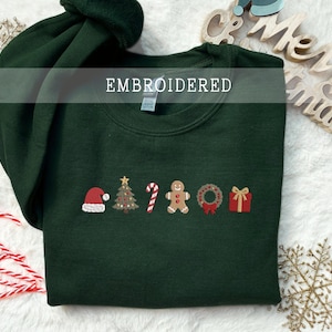 Könnte beinhalten: Ein dunkelgrüner Rundhals-Pullover mit einer gestickten Reihe von Weihnachtsmotiven, darunter eine rote Weihnachtsmannmütze, ein geschmückter Tannenbaum, eine Zuckerstange, ein Lebkuchenmann, ein grüner Kranz mit roter Schleife und ein rotes Geschenk.