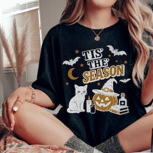 May include: Black t-shirt with a Halloween-themed graphic. The design includes a white cat, a jack-o'-lantern wearing a witch hat, bats, stars, a crescent moon, and the text "Tis the Season" in gold and white.