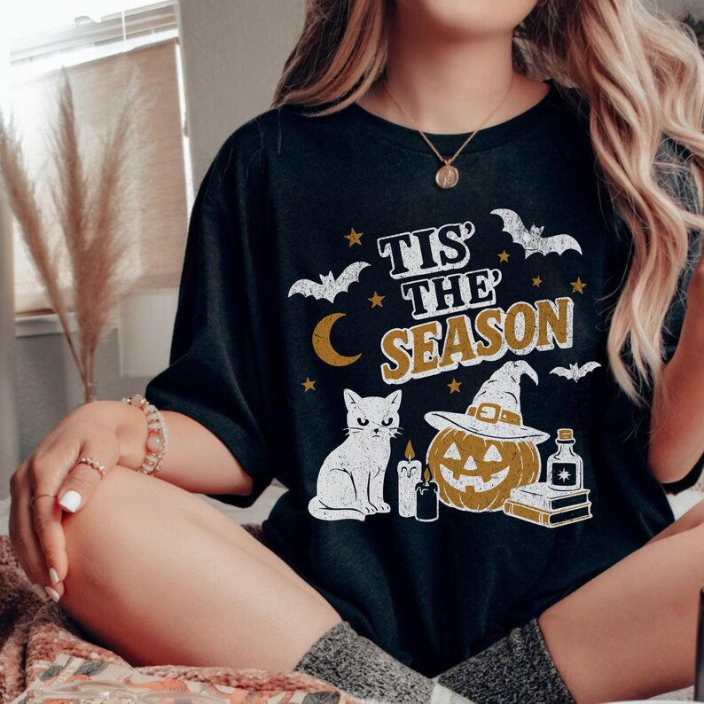 May include: Black t-shirt with a Halloween-themed graphic. The design includes a white cat, a jack-o'-lantern wearing a witch hat, bats, stars, a crescent moon, and the text "Tis the Season" in gold and white.