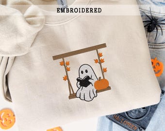 Puede incluir: Sudadera beige bordada con un fantasma leyendo un libro en un columpio con una calabaza. La palabra "EMBROIDERED" está impresa en la parte superior. El diseño incluye detalles marrones, naranjas y negros, perfecto para Halloween.