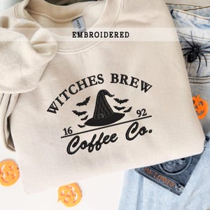 Op de afbeelding: Beige sweatshirt met zwart geborduurde tekst en afbeeldingen. Het ontwerp bevat de woorden "Witches Brew Coffee Co." met een heksenhoed en vleermuizen. Het woord "EMBROIDERED" staat erboven.