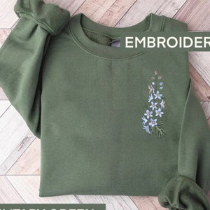 May include: A green sweatshirt with a floral embroidery design. The embroidery features a cluster of white flowers with green leaves. The text "EMBROIDERY" is printed in white on the top right corner of the sweatshirt.