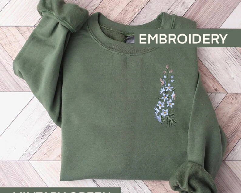 May include: A green sweatshirt with a floral embroidery design. The embroidery features a cluster of white flowers with green leaves. The text "EMBROIDERY" is printed in white on the top right corner of the sweatshirt.