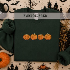 May include: A dark forest green crewneck sweatshirt featuring an embroidered design of four orange pumpkins in a horizontal row across the center. The garment has a ribbed collar, cuffs, and hem, with the word EMBROIDERED printed in a gray bar.